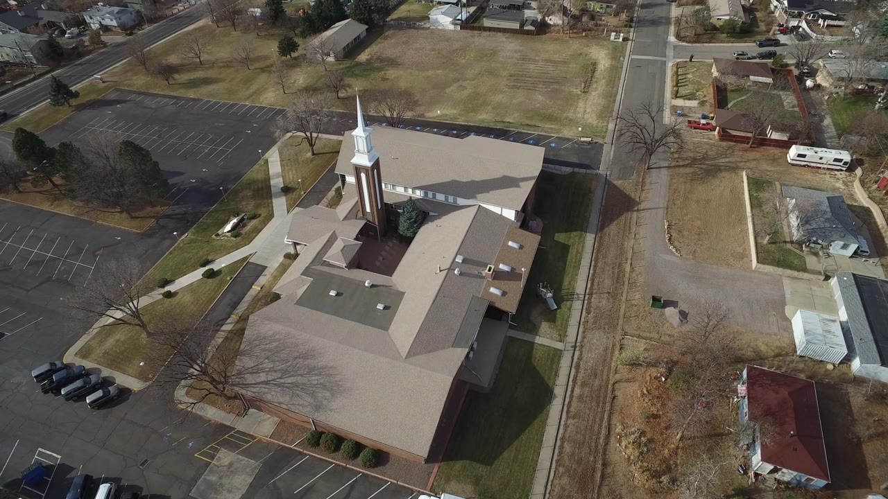 High aerial frame over Westminster, Colorado showing a brick church with a white steeple, tan roofs, empty parking spaces, nearby lawns, roads, and no visible people.