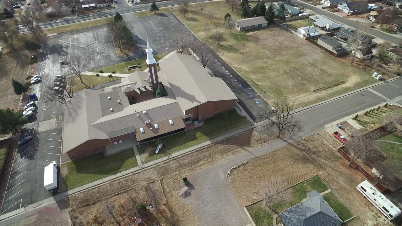 Overhead Westminster, Colorado frame with the church roof and steeple centered, paved parking to the left, houses and trailers beyond the road, and no visible people.