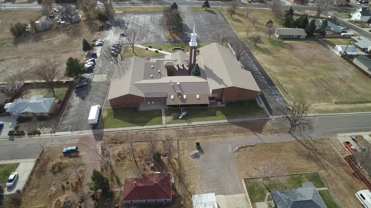 High aerial frame in Westminster, Colorado with the church roof centered, cars along the parking lot edge, surrounding houses, bare trees, and no clearly visible people.