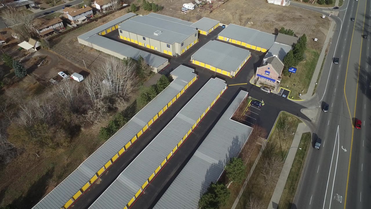 Aerial frame in Westminster, Colorado showing long rows of yellow doors on low buildings, a paved driveway network, vehicles on the adjacent road, and no visible people.