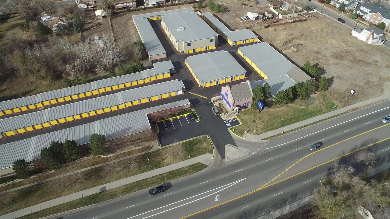 Aerial frame over Westminster, Colorado with the road shifted across the lower foreground, several cars in traffic lanes, yellow-trimmed buildings, open ground, and no visible people.
