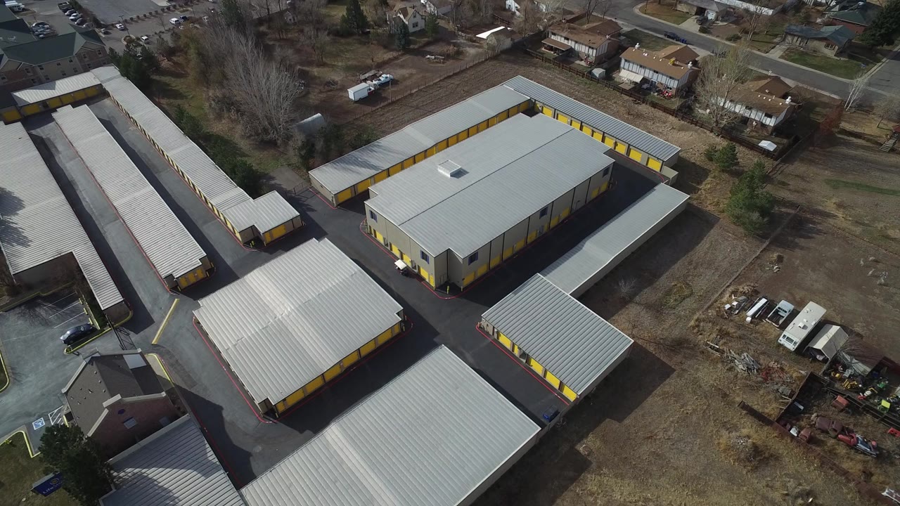 Drone frame looking across the Westminster warehouse roofs and yellow door rows, with residential houses and leafless trees behind the site and no visible people.