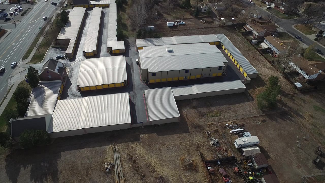 A high-angle aerial frame over Westminster, Colorado shows pale metal-roof industrial buildings, yellow doors, roadside traffic, dirt lots, and no visible people.