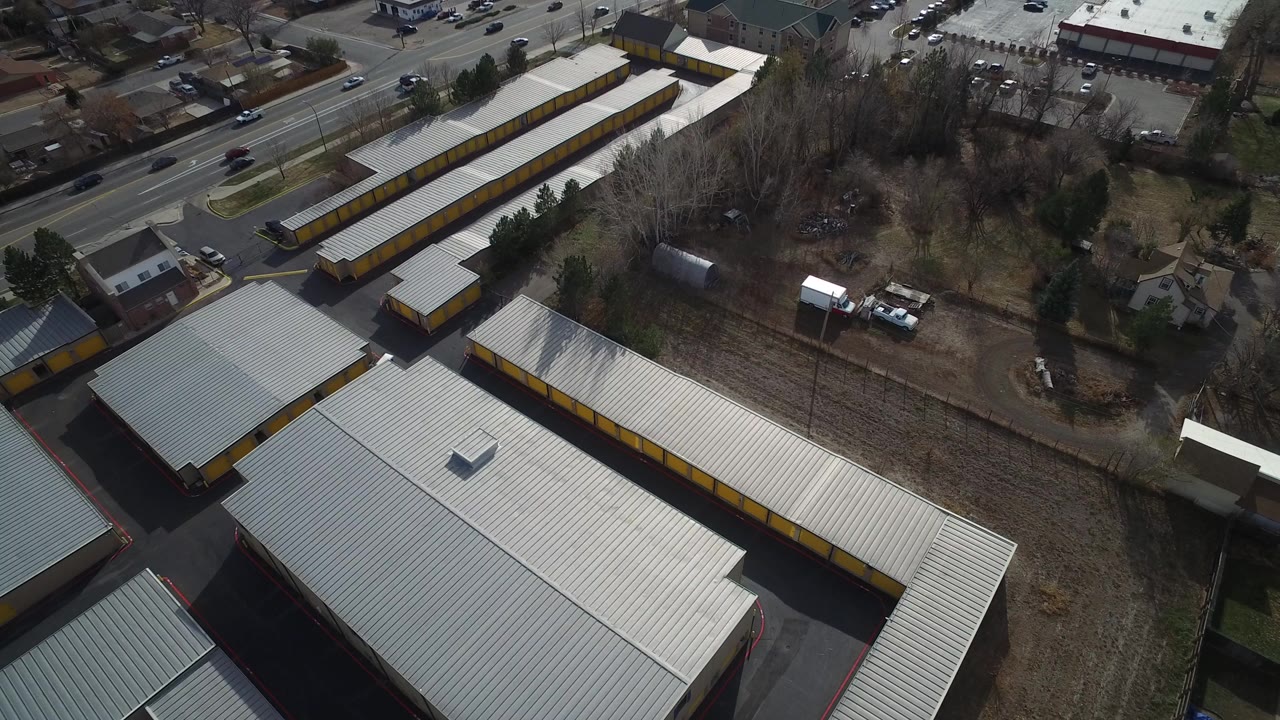 High aerial view in Westminster, Colorado showing white-roofed commercial buildings, yellow trim, a road with vehicles, leafless trees, and no visible people.
