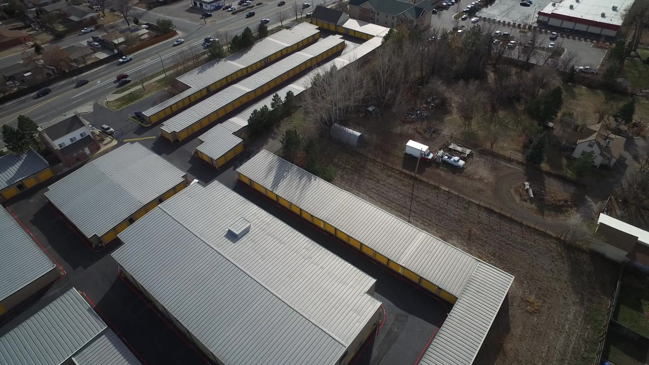 Aerial frame in Westminster, Colorado looking across long white roofs toward a road with moving vehicles, parking areas, trees, and no visible people.