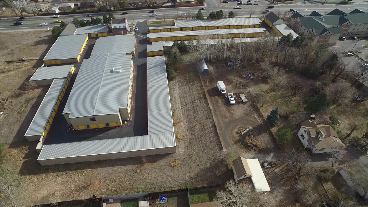 Opening aerial frame in Westminster, Colorado showing a yellow commercial building with a gray roof, surrounding houses, cars on nearby roads, brown grass, bare trees, and no visible people.