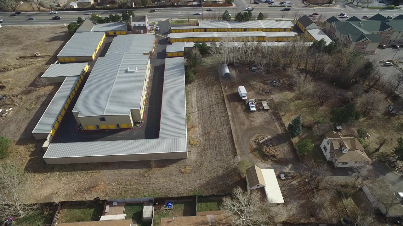 Aerial frame of a gray-roofed yellow building near homes, trees, cars, and a wide road.