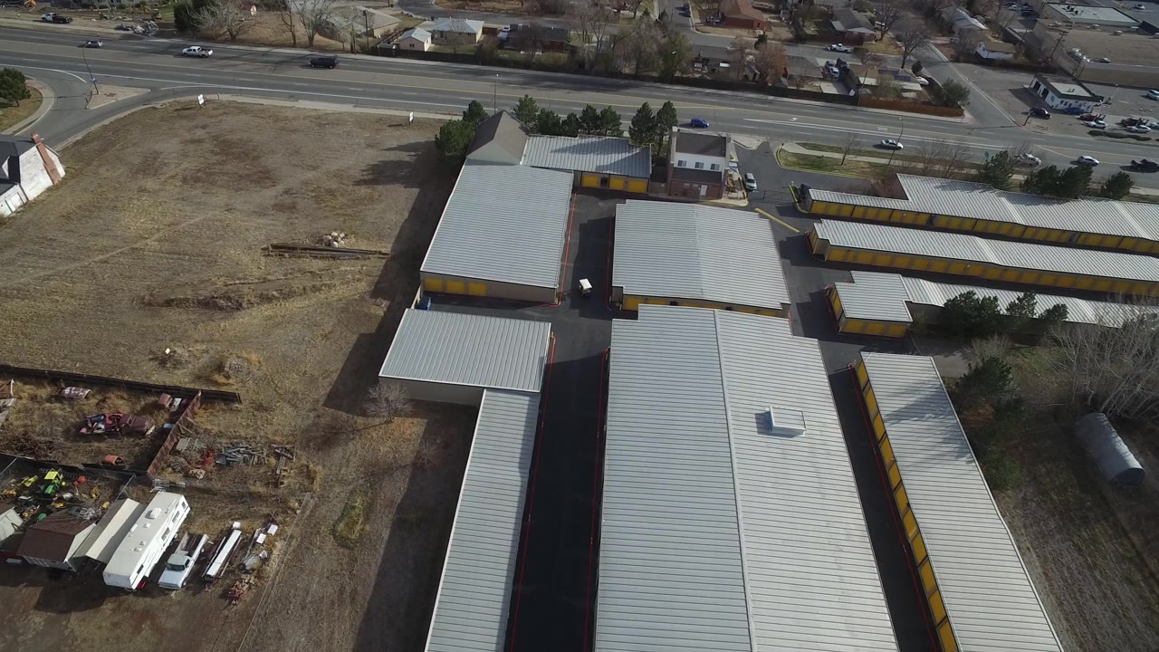 High drone view in Westminster, Colorado with long gray roofs and yellow door rows below a multi-lane road, a vacant lot, and no visible people.