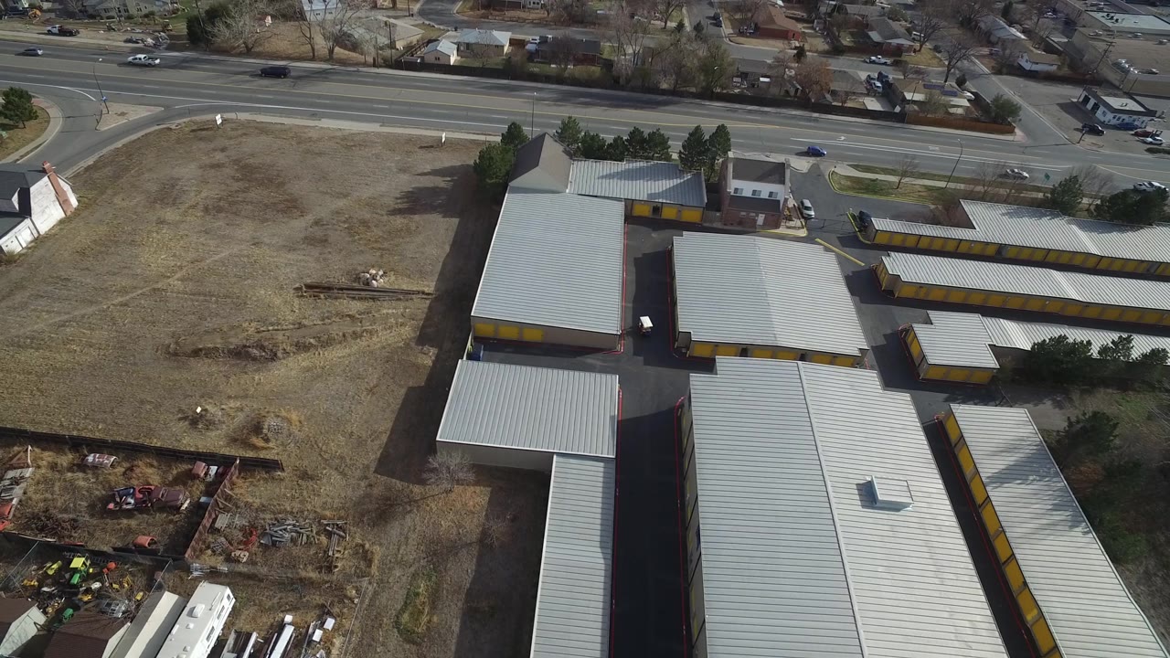 Aerial frame in Westminster, Colorado showing storage-like buildings with gray roofs, a broad dry lot, traffic on the road, homes in the background, and no visible people.