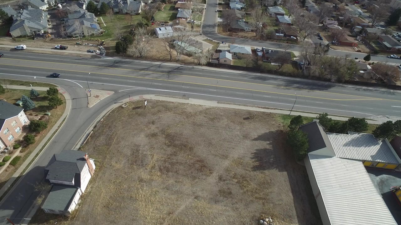 Aerial frame over Westminster, Colorado showing a wide road, dry open lot, nearby homes, sidewalks, and a few cars, with no visible people.