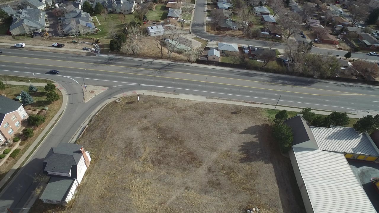 Drone view in Westminster, Colorado with the road running across the frame, houses beyond it, a dry lot below, and no visible people.