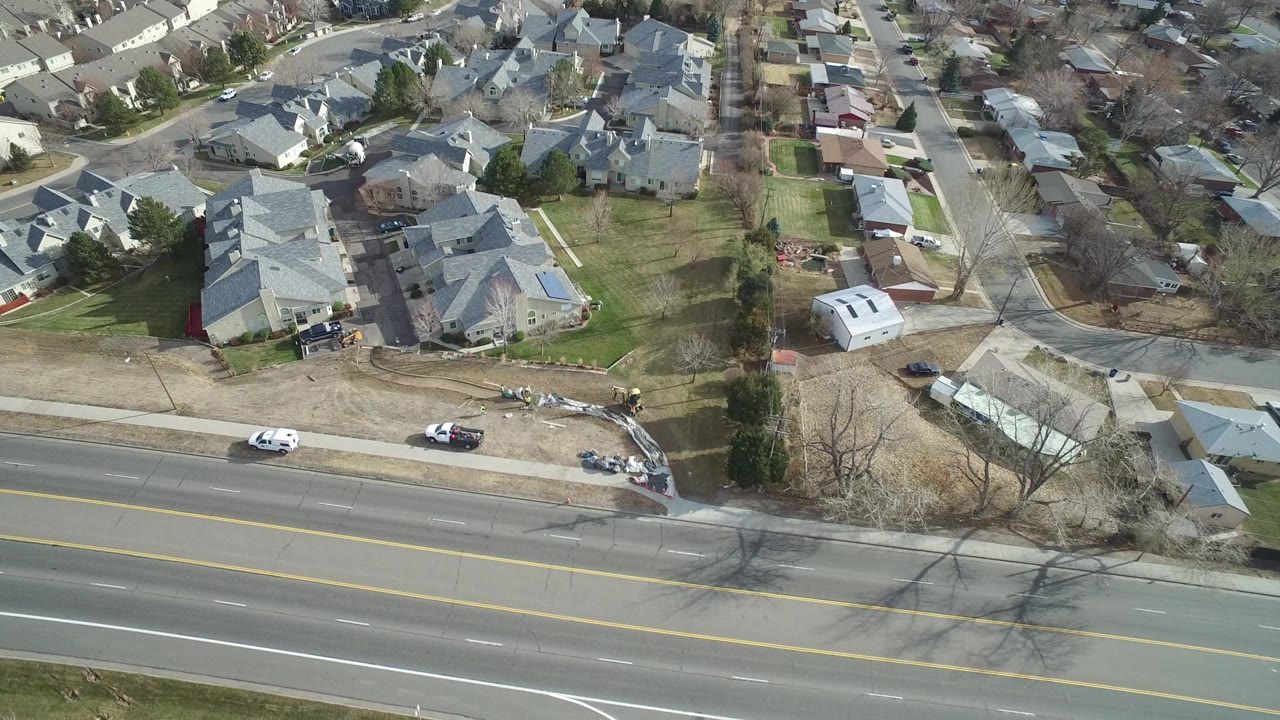 High aerial view over Westminster, Colorado homes with gray roofs, small lawns, a wide road along the bottom edge, parked cars, and no visible people.