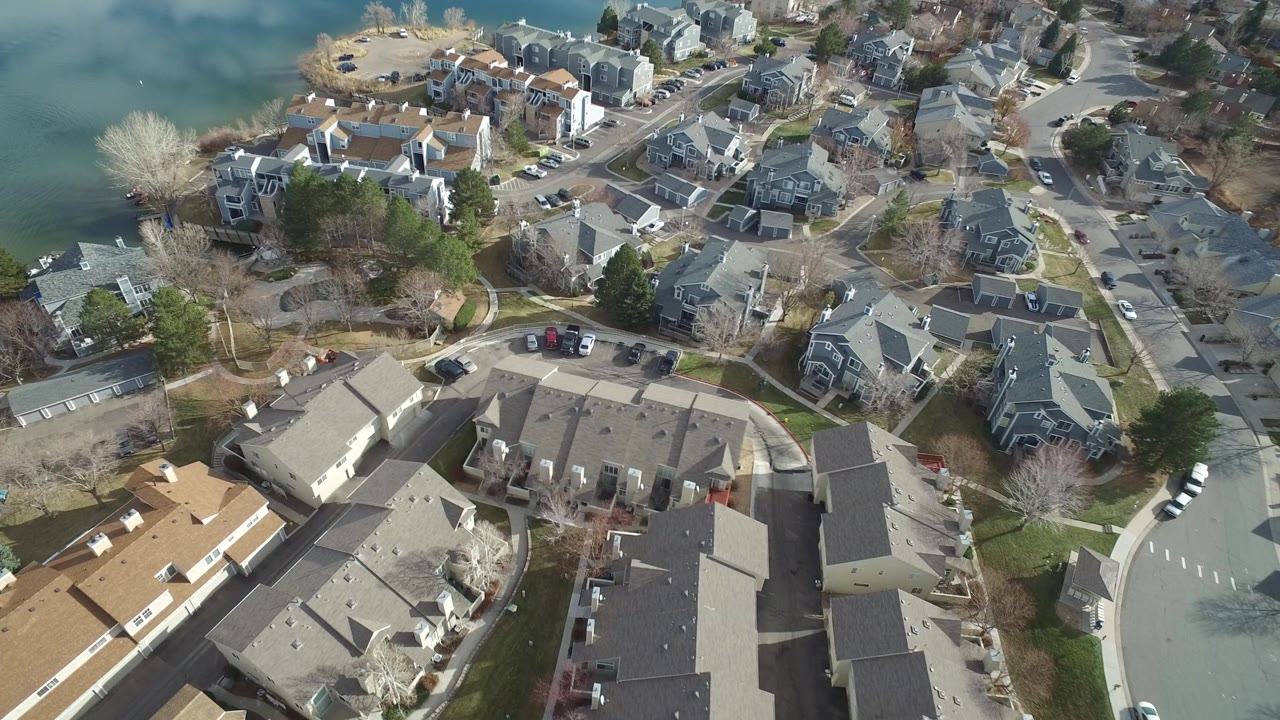 Aerial view over Westminster, Colorado homes near green-blue water, with curved roads, parked cars, leafless trees, and no recognizable people visible.