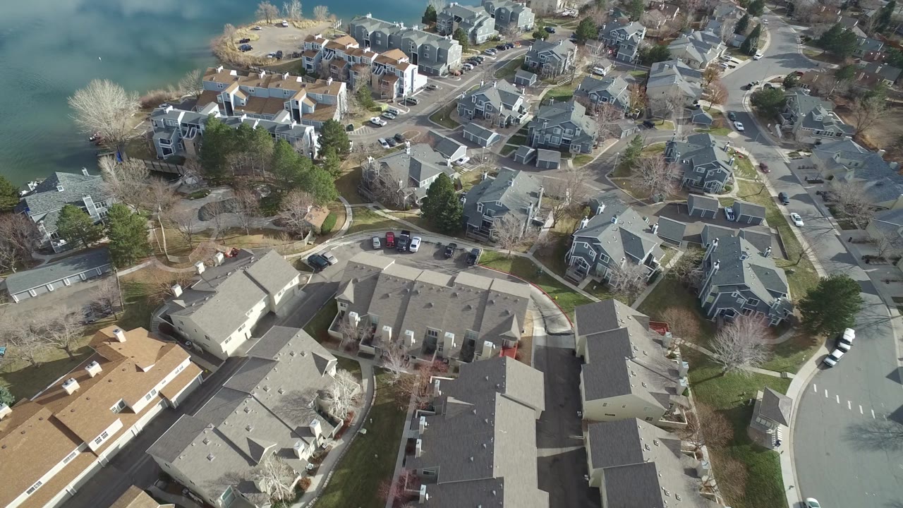 High aerial frame of Westminster, Colorado townhomes and houses beside water, with rooflines, driveways, parked cars, bare trees, and no recognizable people visible.