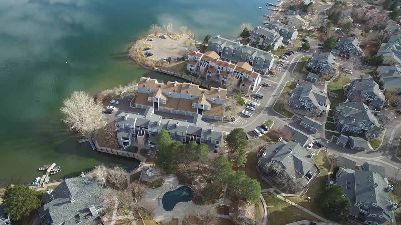 Aerial view of waterfront homes, roads, parked cars, trees, and green water.