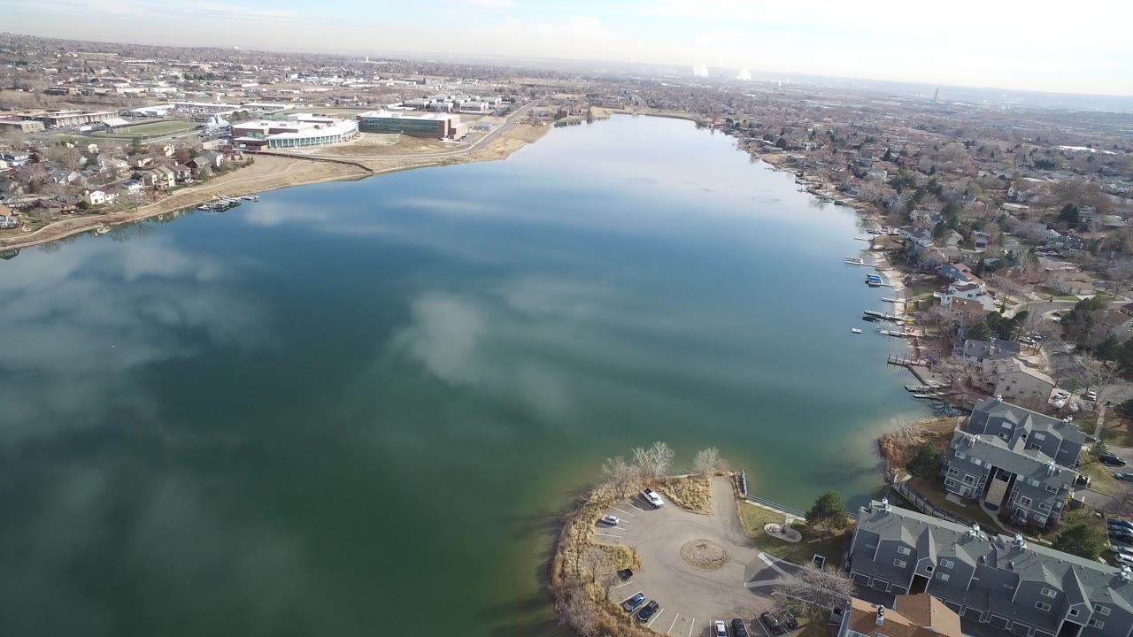 Opening aerial frame over a blue-green lake in Westminster, Colorado, with shoreline homes at the lower right, a large building across the water, distant suburbs, pale sky, and no visible people.