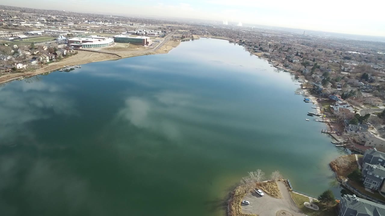 A high aerial frame in Westminster, Colorado shows green-blue water filling most of the view, shoreline homes on the right, a large low building on the far bank, and no visible people.