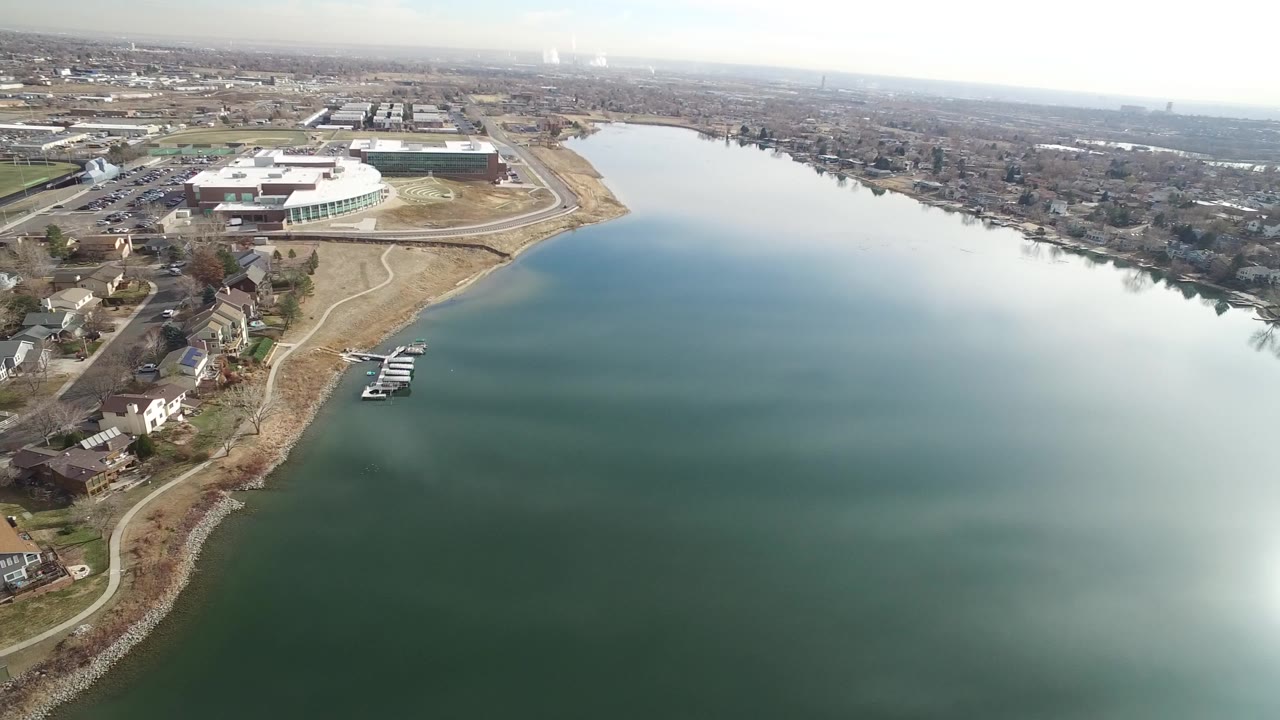 Aerial frame over Westminster, Colorado shows shoreline homes, a small dock, green-blue water, and a large white-roofed structure beyond the shore, with no visible people.