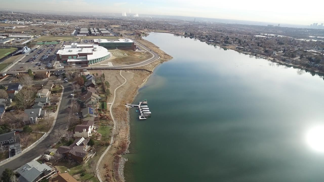 Aerial view over a lake shoreline with homes, docks, and a large white-roofed building.