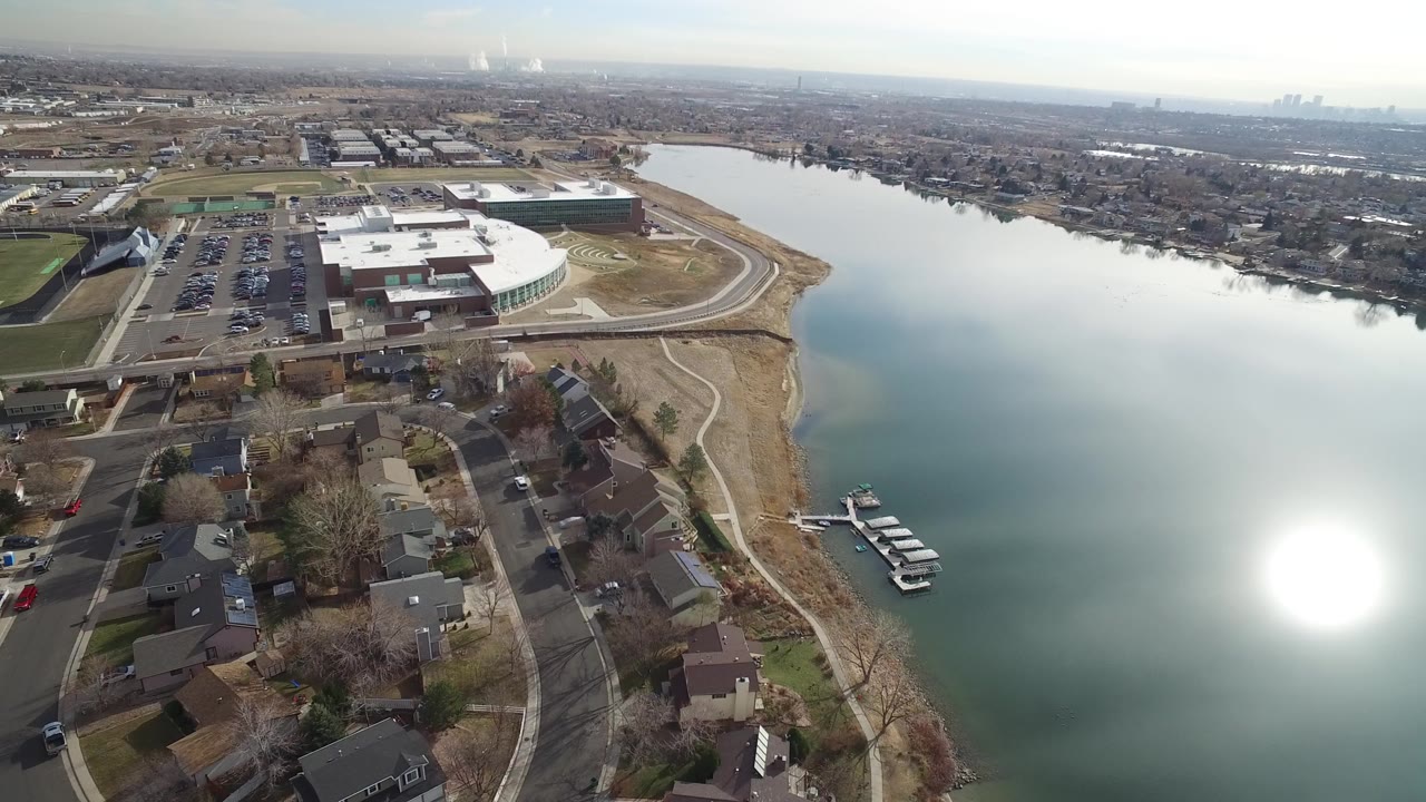 Aerial view of a lake beside houses, roads, a parking lot, and a large curved-roof building.