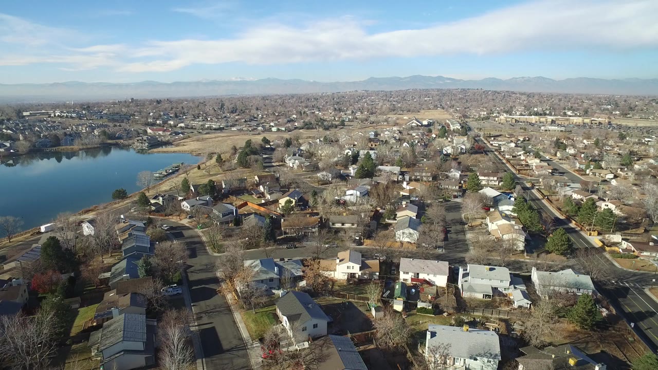 Second aerial frame over Hidden Lake, Colorado, showing more of the lakeshore at left, closely spaced homes below, a main road at right, distant mountains, and no visible people.