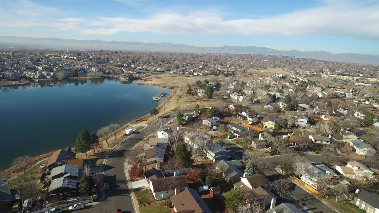 Aerial frame over Hidden Lake, Colorado showing more of the residential blocks around the lake, a shoreline road, clustered rooftops, distant buildings, pale mountains, and no visible people.