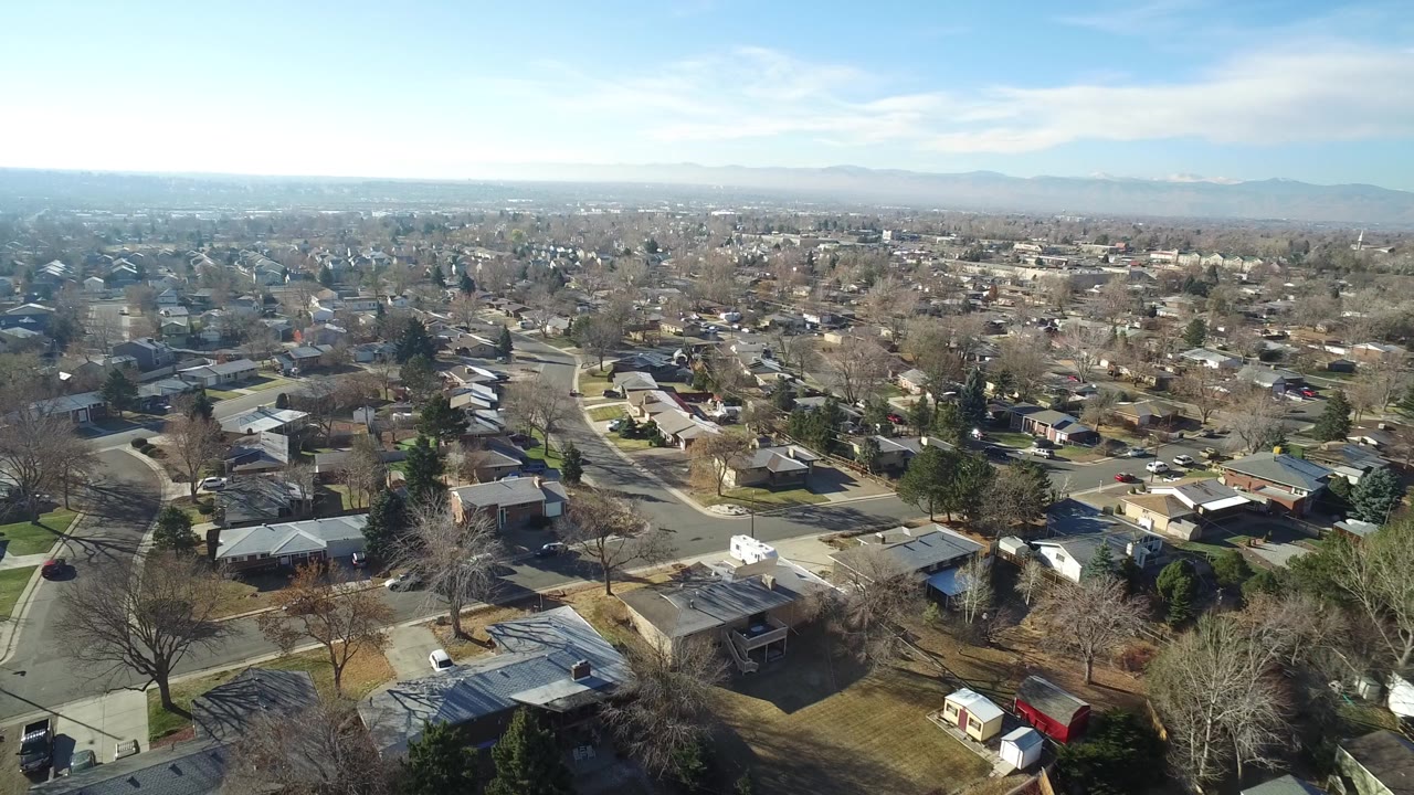Drone-height view across Hidden Lake, Colorado rooftops, intersecting streets, leafless trees, small yards, distant haze, and no visible people.
