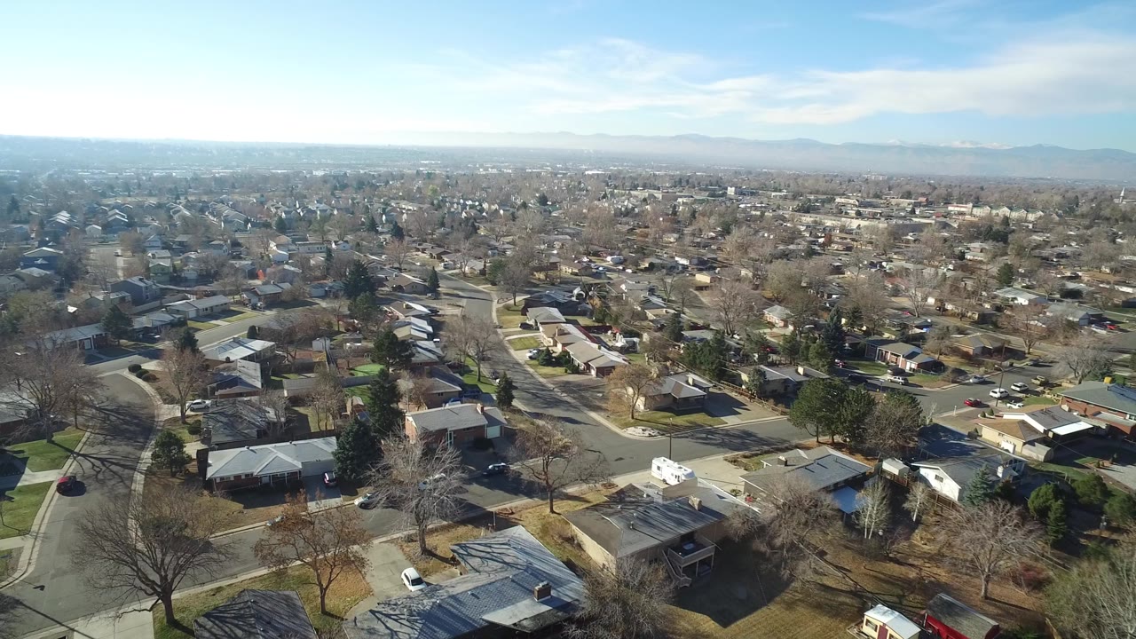 Wide aerial frame in Hidden Lake, Colorado showing suburban houses, branching roads, winter-bare trees, parked cars, distant mountains, and no visible people.