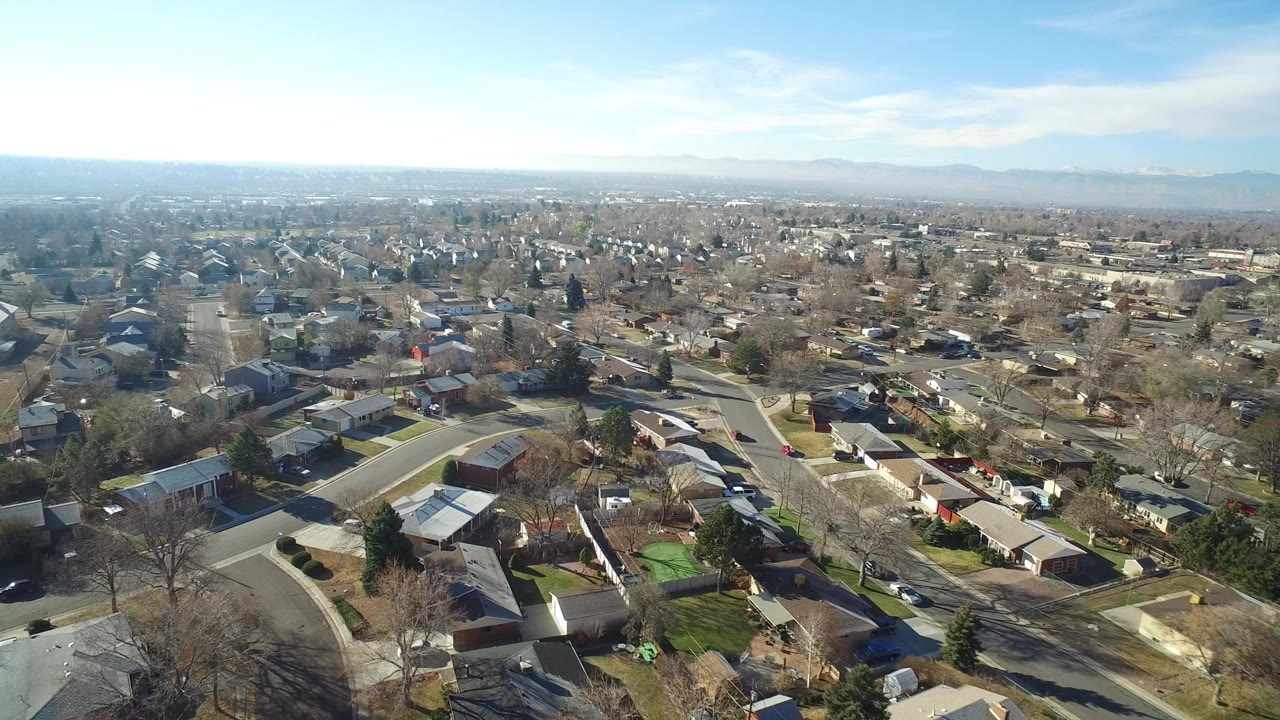 Opening aerial frame over Hidden Lake, Colorado homes, curving residential streets, parked cars, bare trees, and a distant mountain horizon with no visible people.