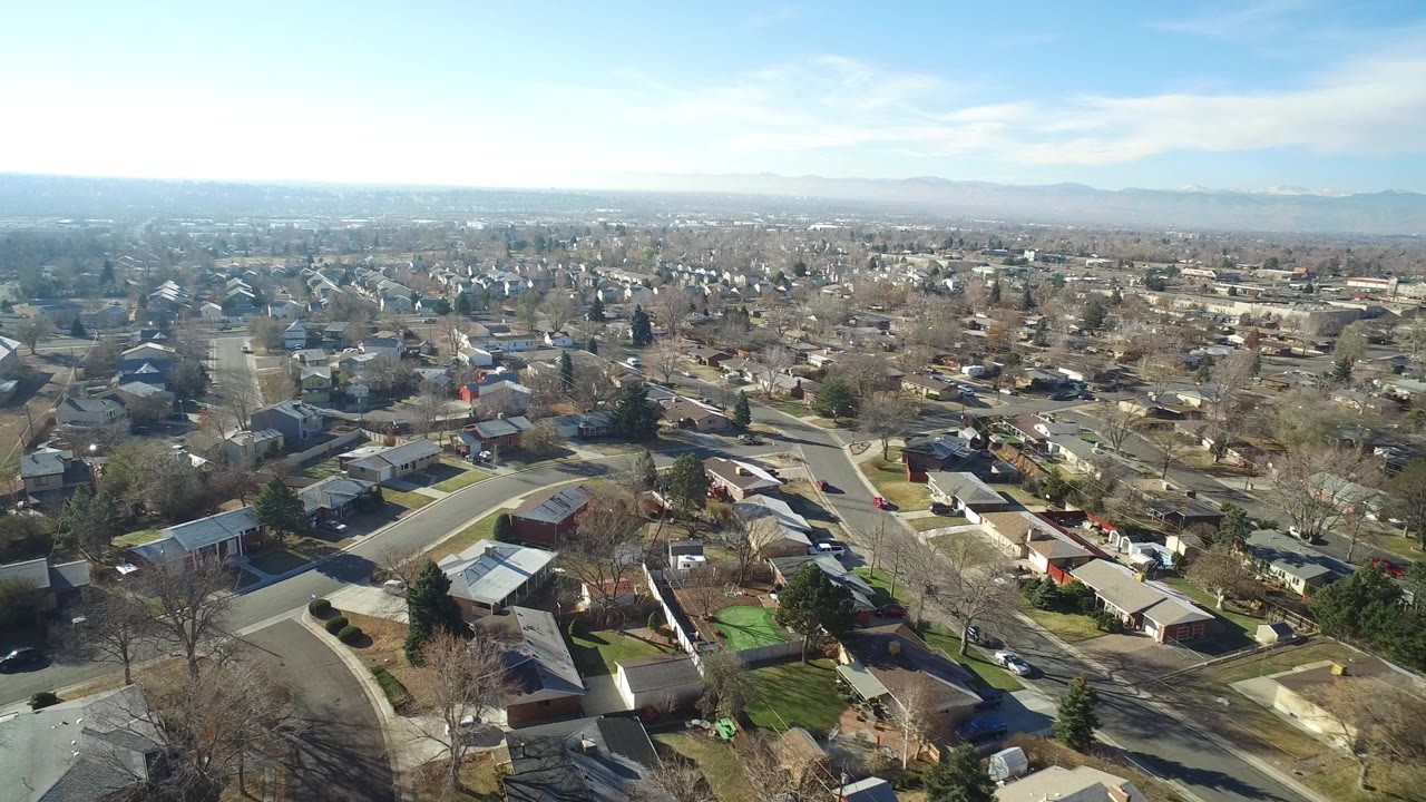 Second aerial frame of Hidden Lake, Colorado showing rooftops, driveways, quiet streets, bare trees, and hazy mountains beyond the neighborhood with no visible people.