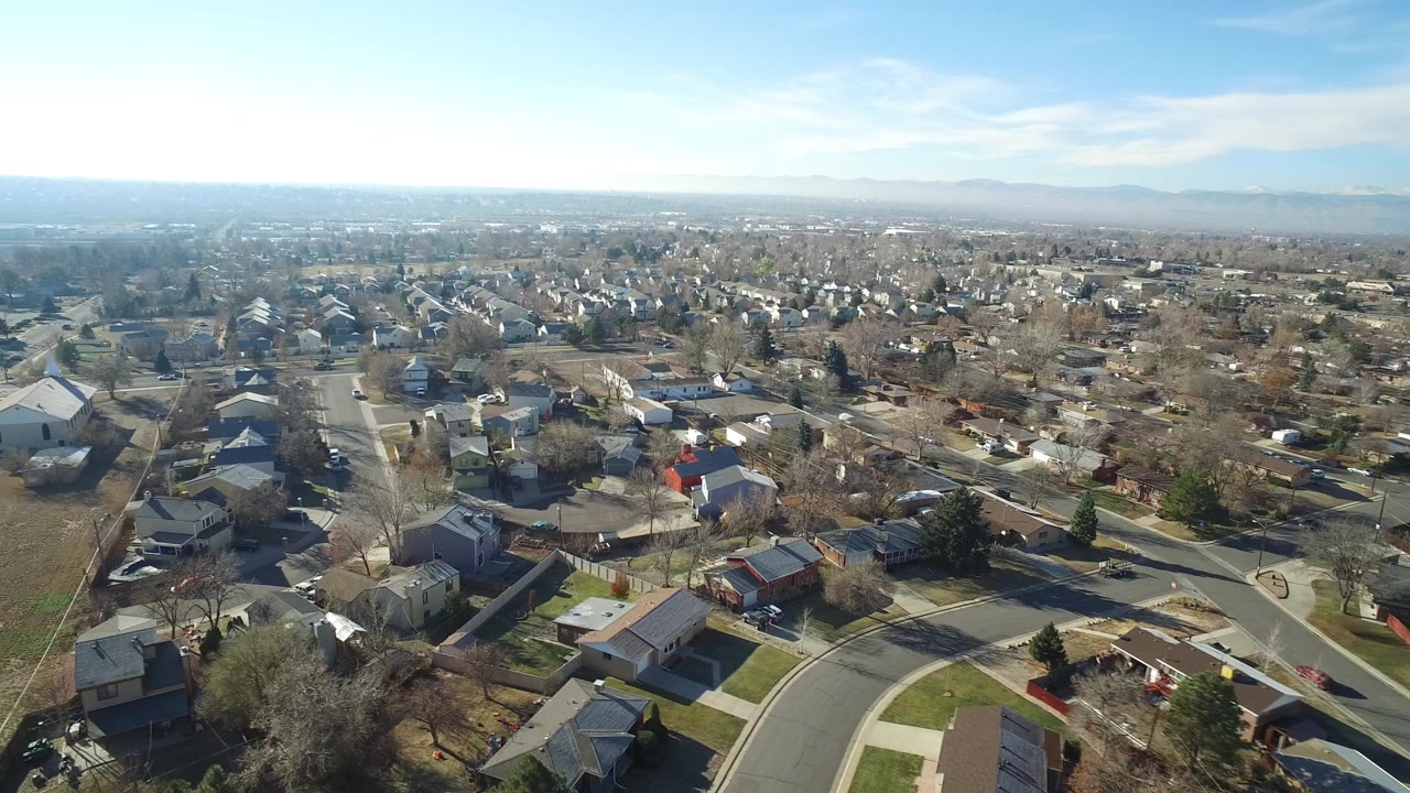 High aerial view over Hidden Lake, Colorado suburban homes, curved streets, leafless trees, and a hazy mountain horizon, with no recognizable people visible.