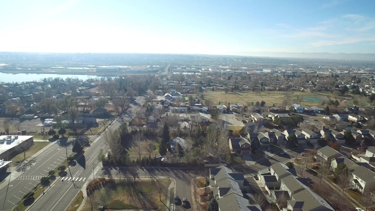 Aerial view over suburban streets, homes, trees, and distant water.