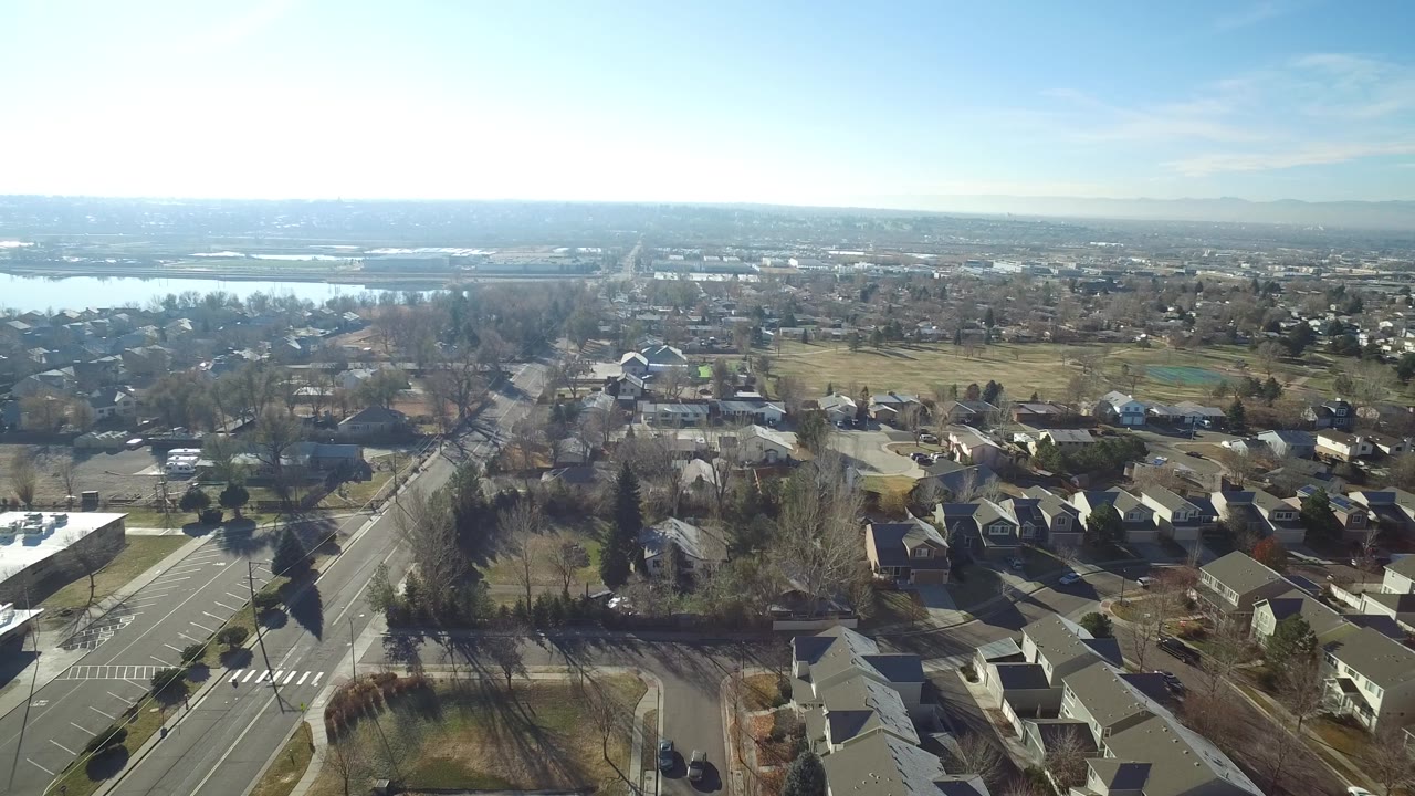 Aerial view in Hidden Lake, Colorado, with rooftops and yards in the foreground, a straight roadway at left, open fields beyond, distant water, and no visible people.