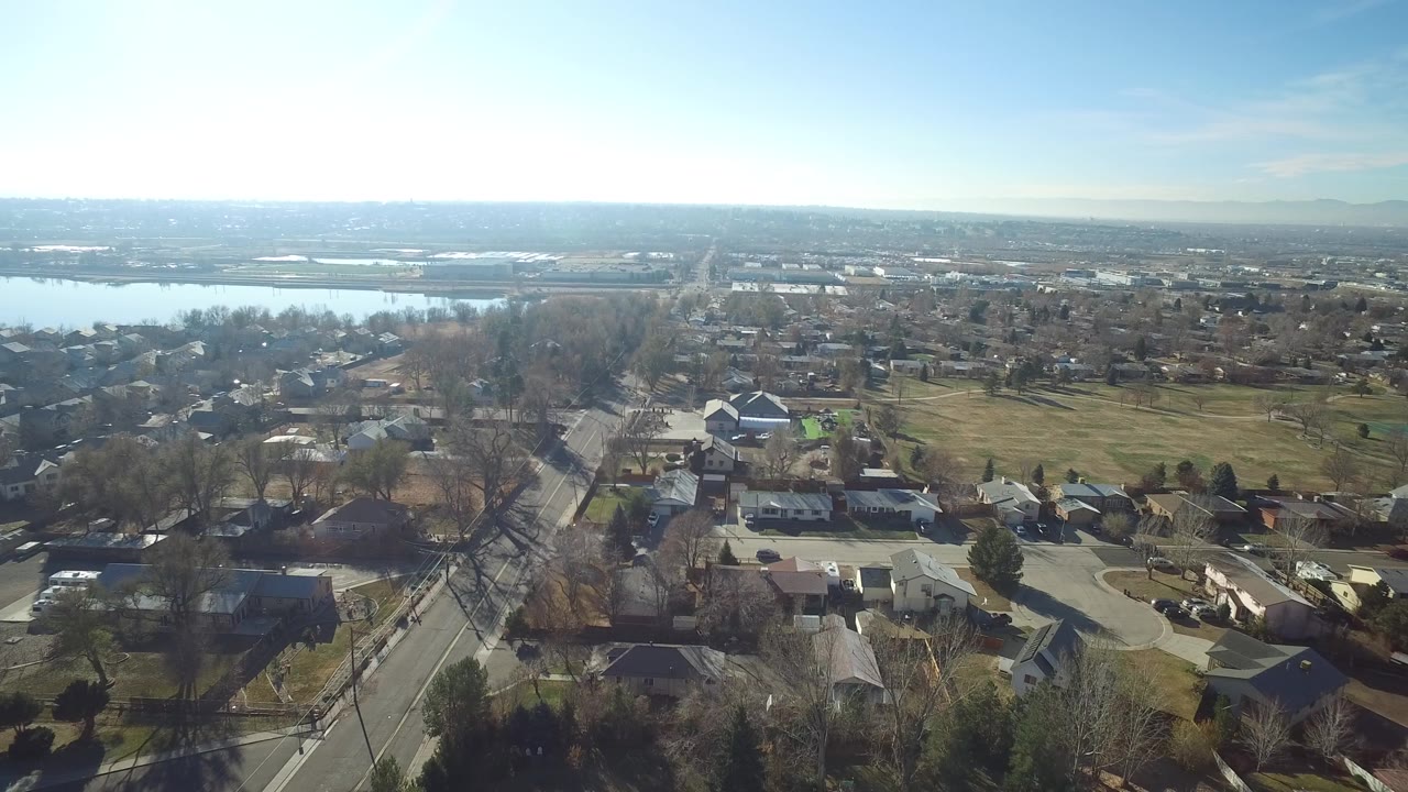 Wide aerial view of houses, streets, trees, open grass, and a lake on the horizon.