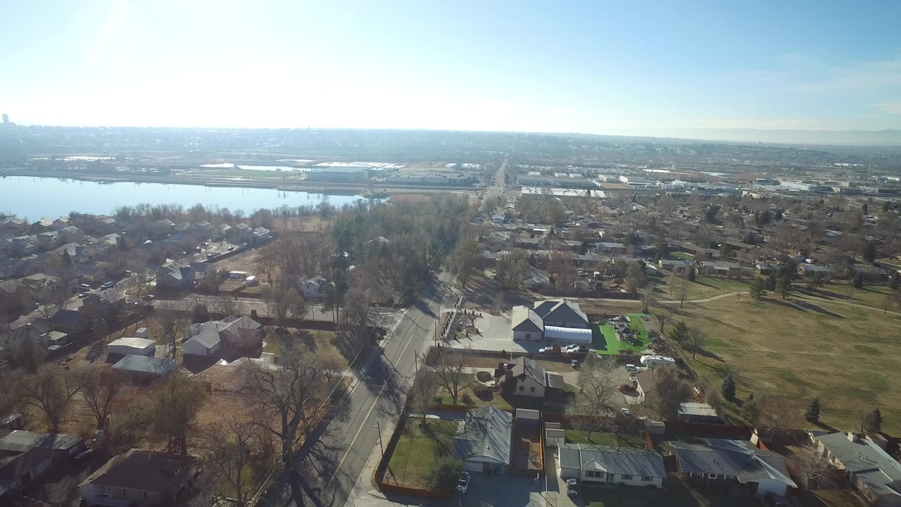 Aerial view of homes, roads, trees, grass, and a lake under a bright sky.