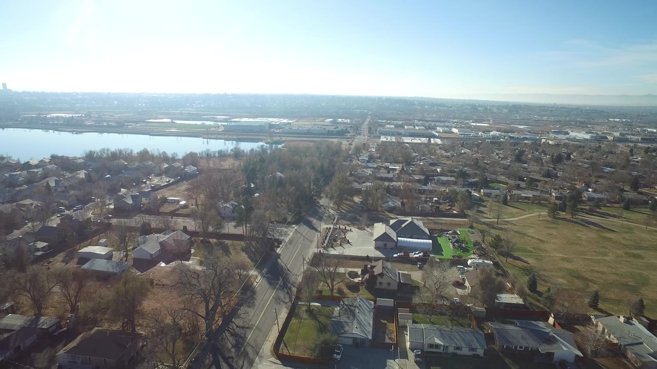 Second frame over Hidden Lake, Colorado, showing a road running through the lower frame, clustered houses, bare trees, a green lawn patch, distant water, and no visible people.