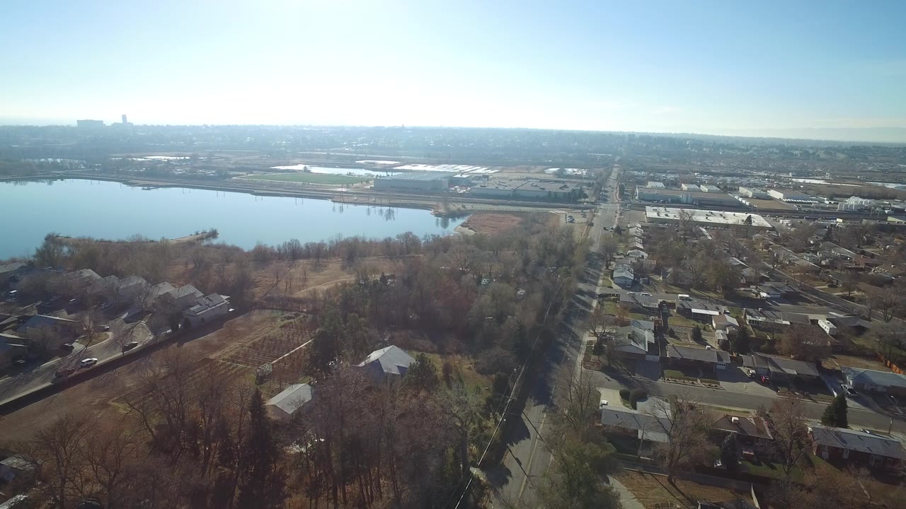 Aerial view of a lake beside houses, trees, roads, and distant industrial buildings.