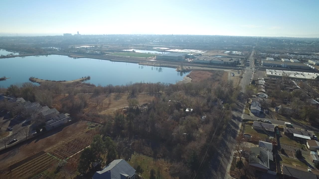 Aerial view of a lake beside trees, roads, homes, and low buildings under a clear sky.