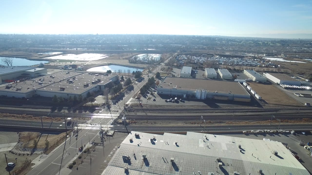 Aerial frame over Hidden Lake, Colorado, centered on a road crossing the rail corridor beside large industrial buildings, reflective ponds, parking areas, and no visible people.