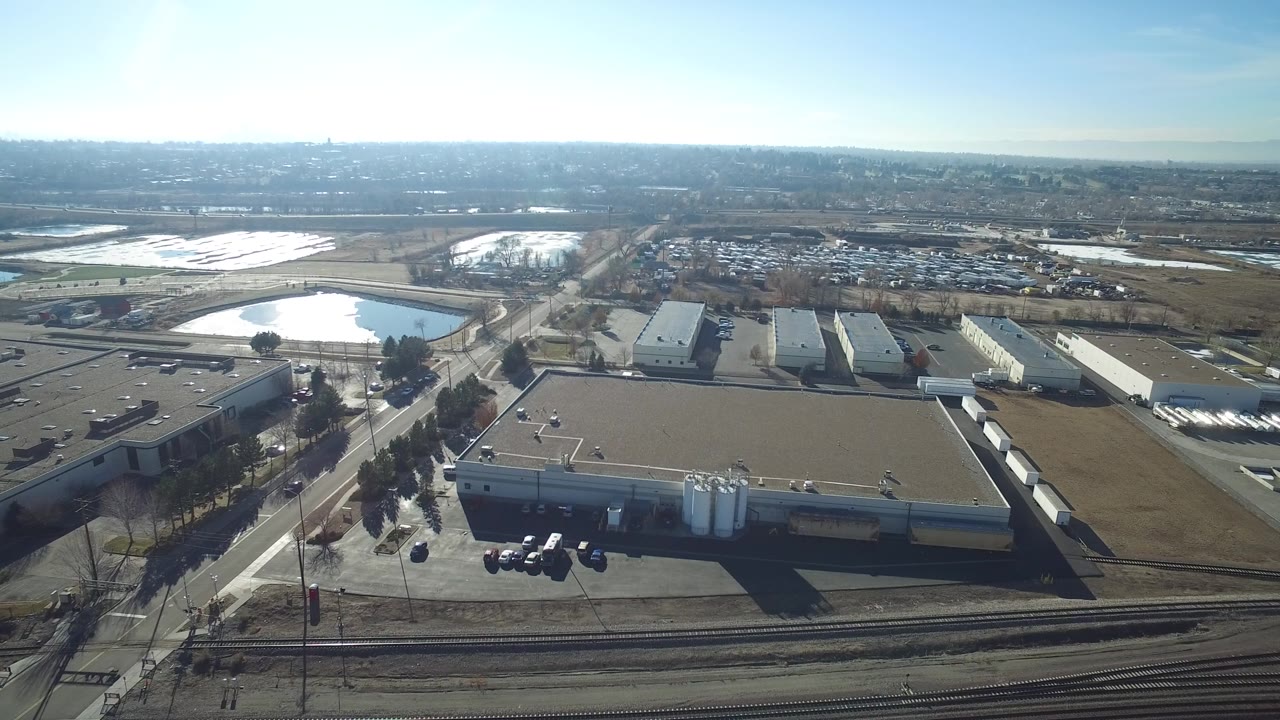 Aerial frame in Hidden Lake, Colorado with a large foreground warehouse roof, white tanks, parked vehicles, the blue lake to the left, and no visible people.