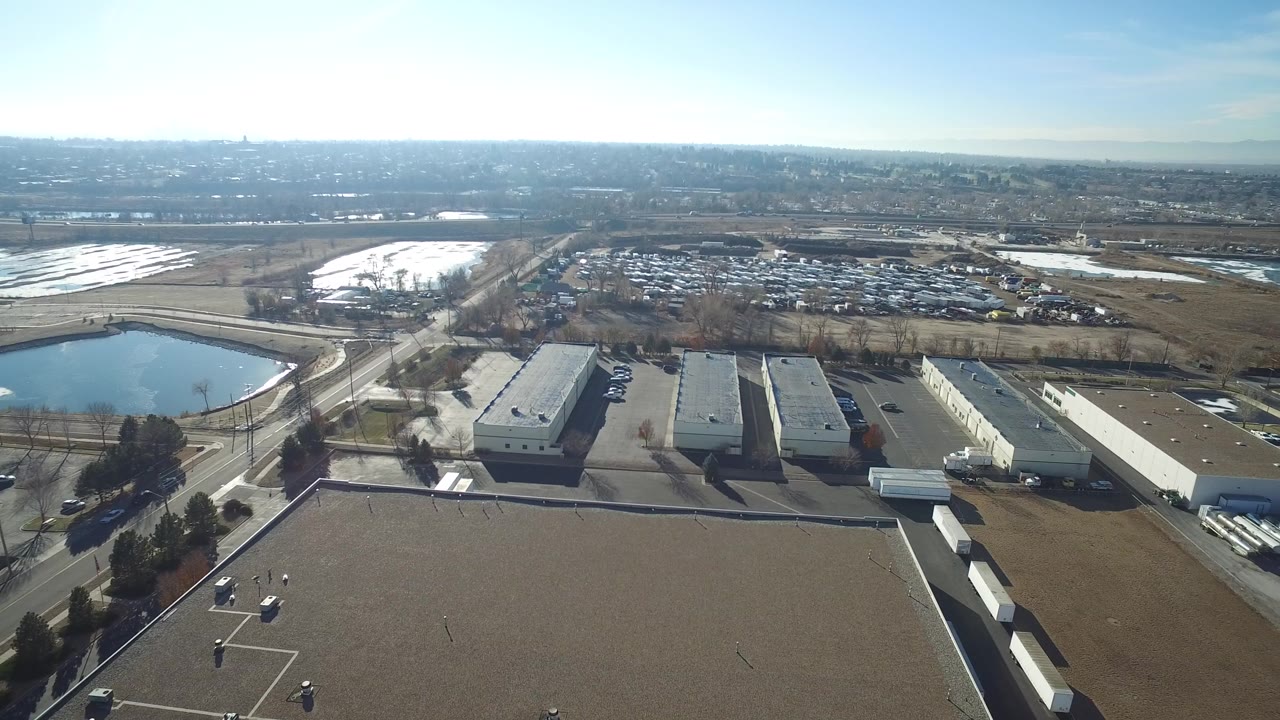 Aerial frame over Hidden Lake, Colorado showing flat industrial rooftops, a small pond, roads, open fields, distant neighborhoods, and no visible people.