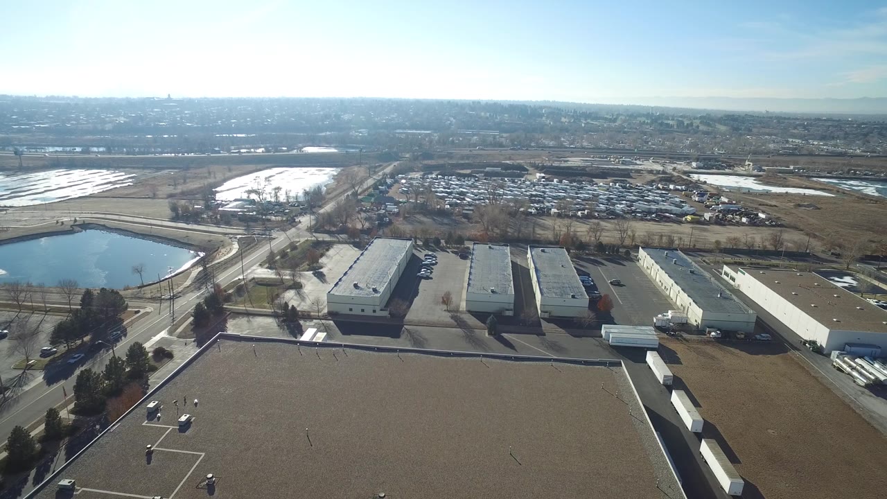 Drone-height view in Hidden Lake, Colorado with warehouse roofs in the lower frame, a pond at left, brown fields beyond, parked vehicles, and no visible people.