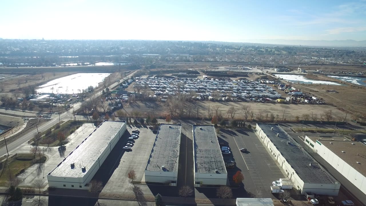 Wide aerial view over Hidden Lake, Colorado, with flat-roofed commercial buildings in the foreground, a large vehicle lot and open fields beyond, bright reflective ground at left, and no visible people.