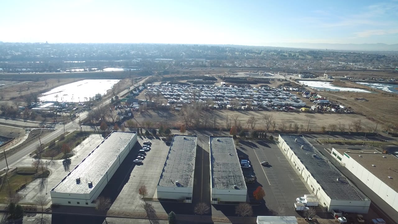 Aerial frame in Hidden Lake, Colorado showing warehouse-style roofs at the lower edge, parking rows beside them, a diagonal road, a large vehicle lot, dry fields, and no visible people.