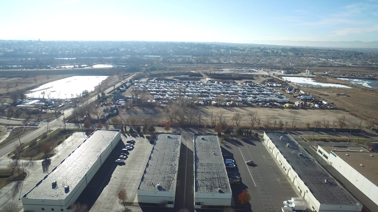 High aerial view of parking lots, low commercial buildings, roadside trees, open land, and distant neighborhoods.