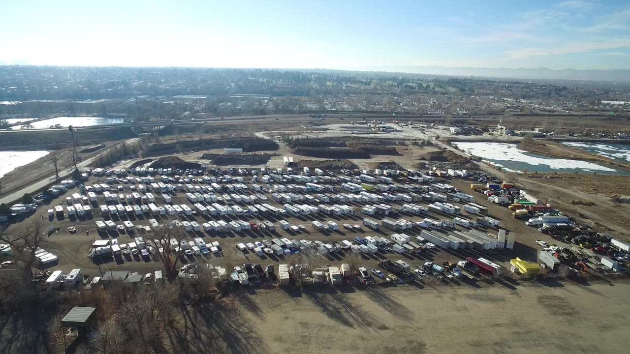 Second frame in Hidden Lake, Colorado, looking across dense rows of parked cars toward water, roads, distant low buildings, bright sky, and no visible people.
