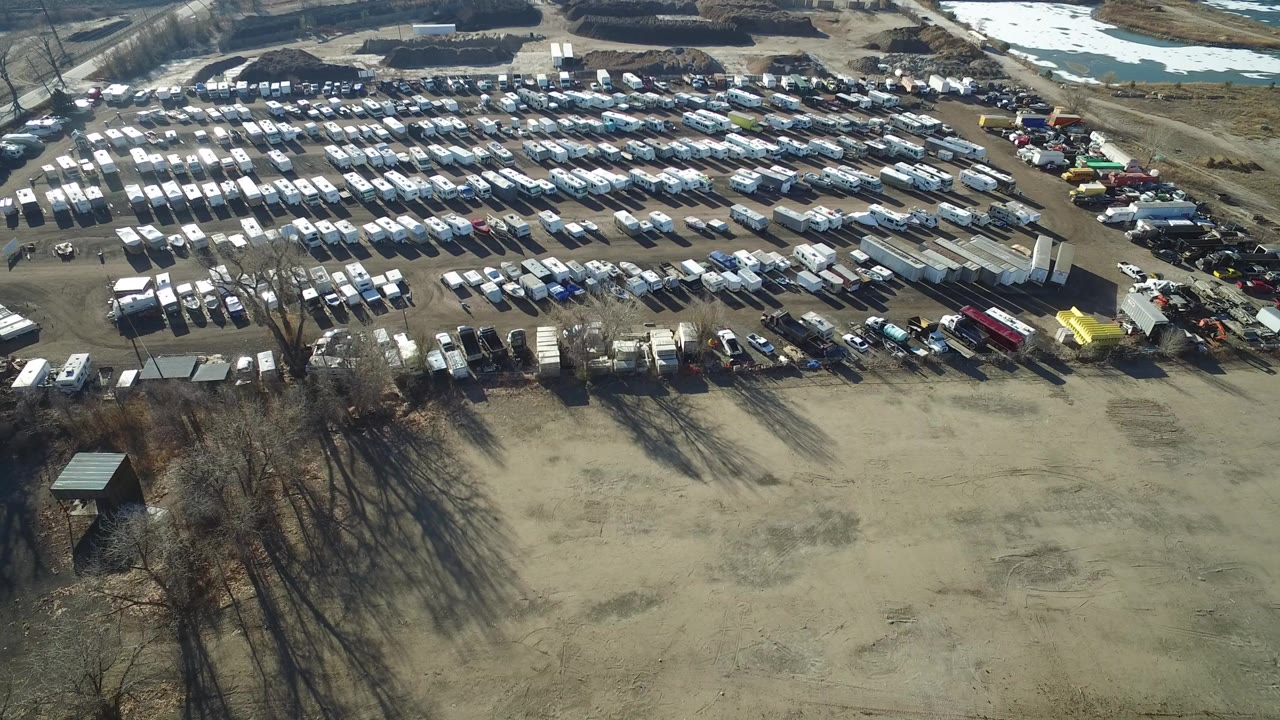 Opening aerial frame in Hidden Lake, Colorado, showing an empty dirt foreground, bare trees, dense rows of parked vehicles, and no visible people.