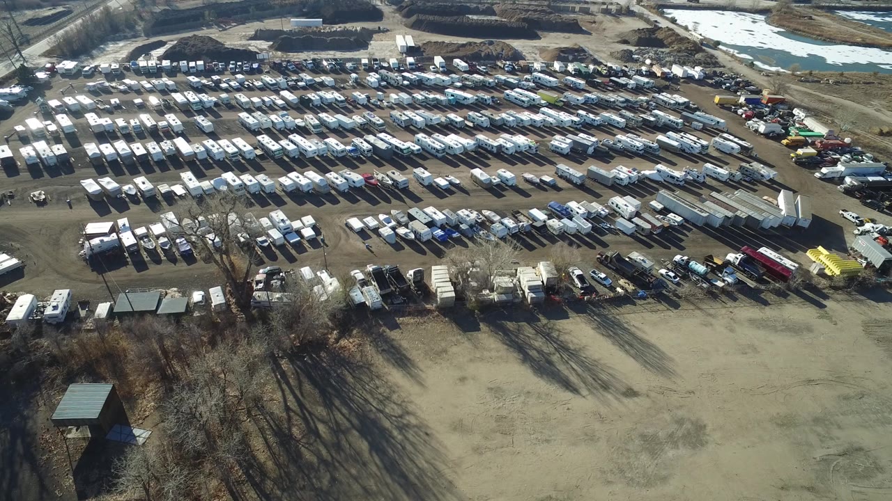 Second frame in Hidden Lake, Colorado holds the oblique aerial view across dense rows of white vehicles, foreground trees, dirt access roads, and distant open ground, with no visible people.