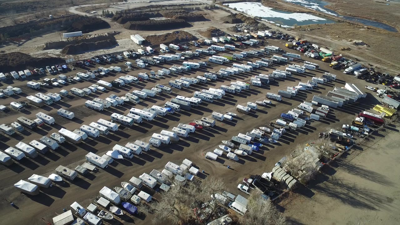 Opening overhead frame of a large Hidden Lake, Colorado vehicle yard with rows of RVs and trailers, open brown land beyond, snow patches near water, and no visible people.