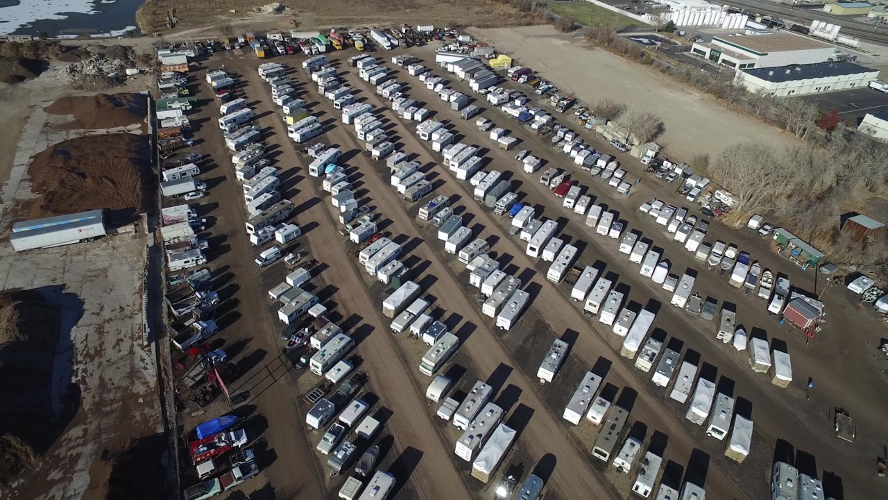 Aerial view in Hidden Lake, Colorado, with the packed parking rows centered more tightly and a dirt access road and pale industrial roofs visible beyond the lot.